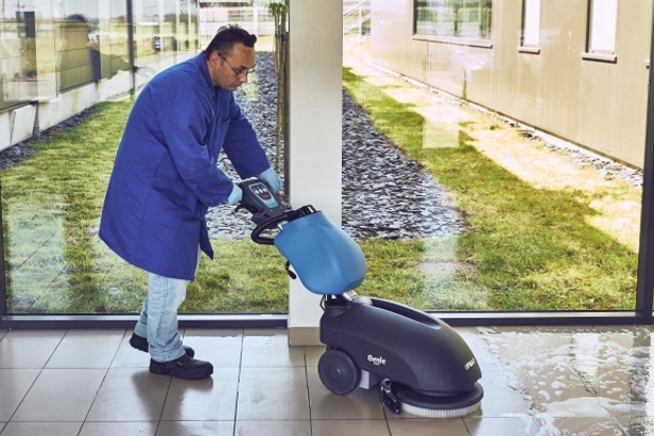Homme portant une blouse bleue et des gants de protection utilisant une autolaveuse dans un hall vitré. De grandes baies vitrées donnent sur des espaces verts à l’extérieur.
