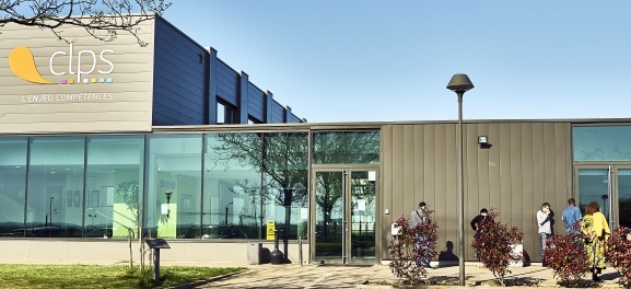 Bâtiment moderne du CLPS avec façade grise, grandes baies vitrées et logo « clps – L’enjeu compétences ». Plusieurs personnes circulent devant l’entrée principale bordée de massifs végétalisés.