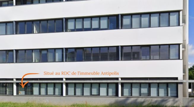 Vue extérieure d'un immeuble de bureaux blanc à plusieurs étages. Une flèche orange indique une fenêtre située au rez-de-chaussée.