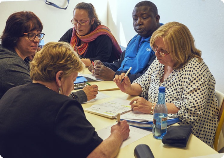 Cinq personnes sont réunies autour d’une table de travail. Plusieurs participants prennent des notes sur des documents tandis que d’autres consultent des supports papier disposés au centre de la table.