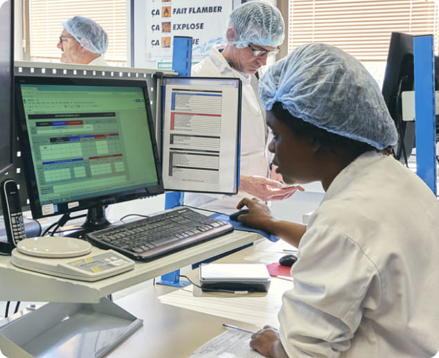 Trois personnes portent des blouses blanches, des charlottes et des lunettes de protection dans un environnement industriel. Au premier plan, une femme utilise un ordinateur équipé d’un écran, d’un clavier et d’une souris, tandis que deux autres personnes consultent des documents à l’arrière-plan.