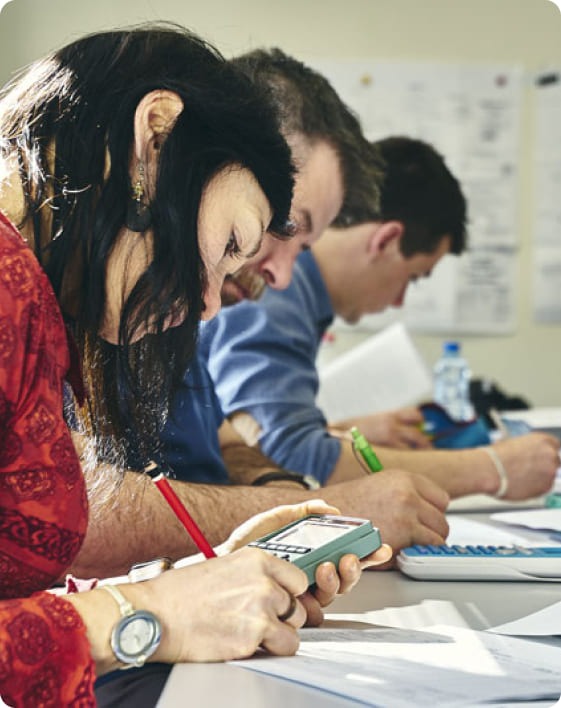 Trois personnes sont assises côte à côte à une table de travail. Au premier plan, une femme tient une calculatrice grise et écrit avec un crayon rouge sur un document. Deux autres personnes écrivent sur des feuilles à l’aide de stylos verts et bleus.