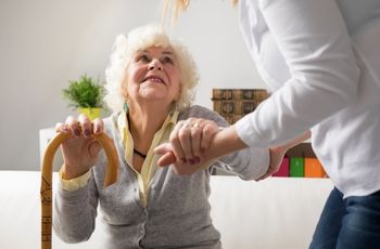 Femme âgée tenant une canne tandis qu'une autre femme l'aide à se lever en lui tenant la main.