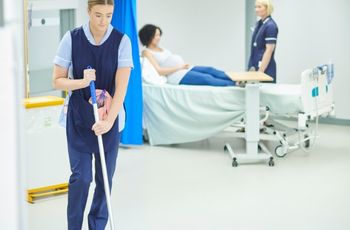Femme en tenue professionnelle réalisant le nettoyage d’un sol dans une chambre équipée d’un lit médicalisé.