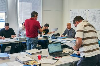 Personnes participant à une formation autour de tables couvertes de documents, tandis qu'un formateur debout présente une feuille au groupe.
