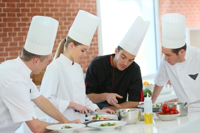 Quatre personnes en tenue de cuisine travaillent autour d’un plan de travail. Au centre, un homme en veste noire montre un geste technique à une femme en tenue blanche qui découpe des ingrédients. Des assiettes, des bols, une bouteille d’huile et des légumes sont disposés sur le plan de travail.