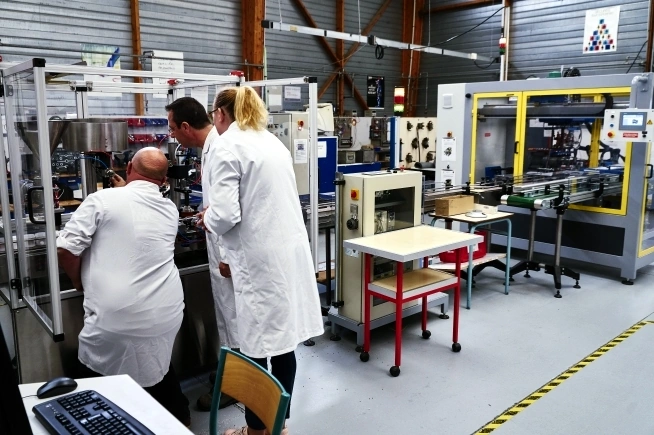 Trois personnes portant des blouses blanches interviennent sur une ligne de production industrielle automatisée. Deux personnes observent l’intérieur d’une machine protégée par des parois transparentes tandis qu’une troisième personne se tient à leurs côtés. Des convoyeurs, postes de commande et équipements industriels occupent l’ensemble de l’atelier.