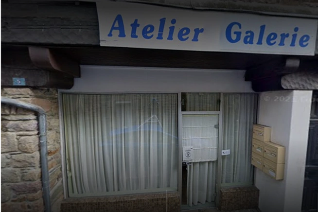 Façade d’un local avec une grande vitrine protégée par des rideaux verticaux clairs. Une enseigne blanche portant l’inscription « Atelier Galerie » en lettres bleues est installée au-dessus de l’entrée. Des boîtes aux lettres sont fixées à droite de la porte.