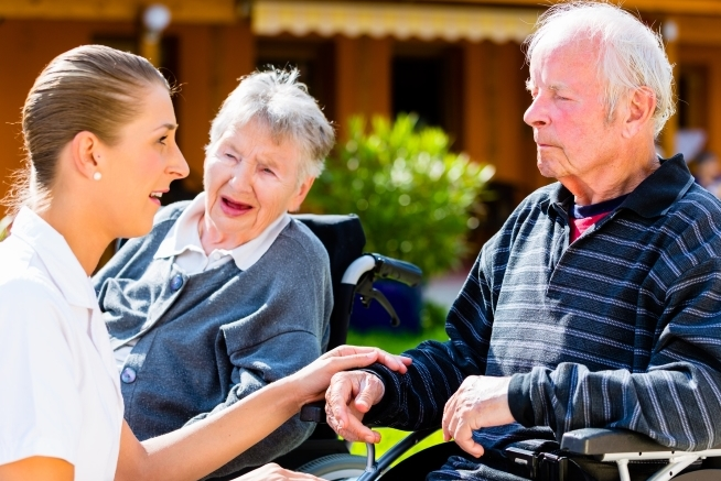 Trois personnes sont réunies en extérieur autour de deux personnes âgées utilisant un fauteuil roulant. Une femme en tenue claire est assise à côté d'un homme âgé en fauteuil roulant et lui tient la main. Une femme âgée en fauteuil roulant est visible à l'arrière-plan.