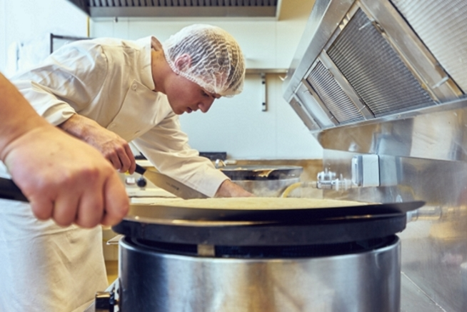 Personne en tenue de cuisine blanche et charlotte manipulant une grande spatule sur une billig. Au premier plan, une plaque de cuisson ronde occupe le centre de l’image tandis qu’une autre main tient un ustensile à gauche.