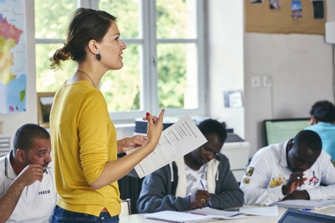 Femme portant un pull jaune et tenant une feuille de travail devant plusieurs participants assis autour d’une table dans une salle de formation. Des cahiers, documents et stylos sont disposés devant les participants.