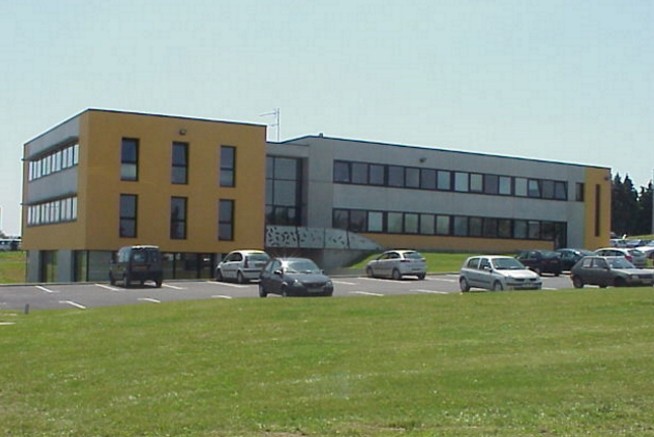 Bâtiment moderne de deux étages aux façades grises et jaunes, avec de larges bandes de fenêtres sur toute la longueur. Plusieurs voitures sont stationnées devant le bâtiment, séparé de la route par une vaste pelouse.