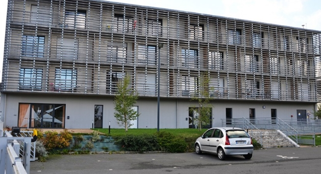 Bâtiment moderne de quatre niveaux avec une façade recouverte de lames horizontales métalliques. Plusieurs fenêtres sont réparties sur l’ensemble de la façade. Un parking, des espaces verts et une voiture stationnée occupent le premier plan.