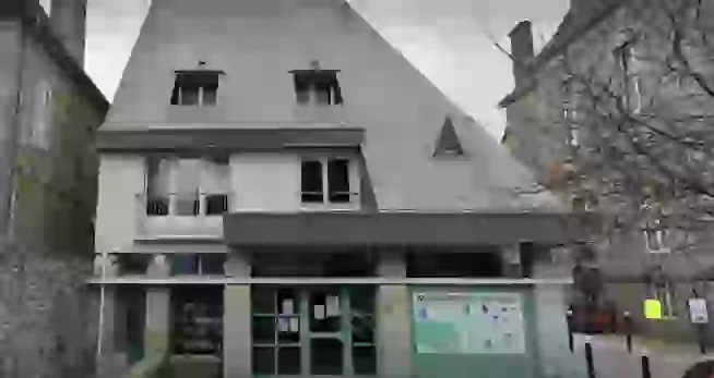 Bâtiment à plusieurs niveaux avec façade claire, grande toiture en ardoise et porche couvert au rez-de-chaussée. Des panneaux d’information sont affichés sur la façade vitrée et un espace piétonnier se situe devant l’entrée.
