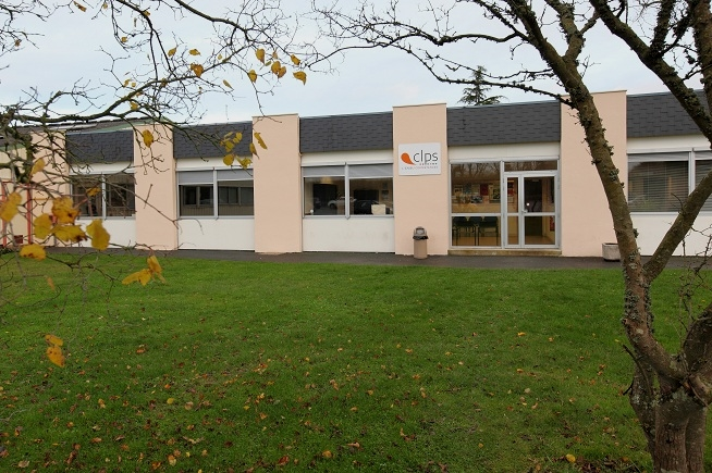Bâtiment de plain-pied aux façades blanches et beige clair portant le logo du CLPS. Une pelouse occupe le premier plan et des arbres encadrent le bâtiment de chaque côté de l’image.