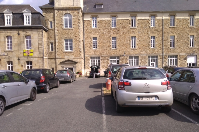 Bâtiment en pierre de plusieurs niveaux avec toiture en ardoise et nombreuses fenêtres blanches. Un parking avec plusieurs véhicules occupe le premier plan, devant la façade principale du bâtiment.