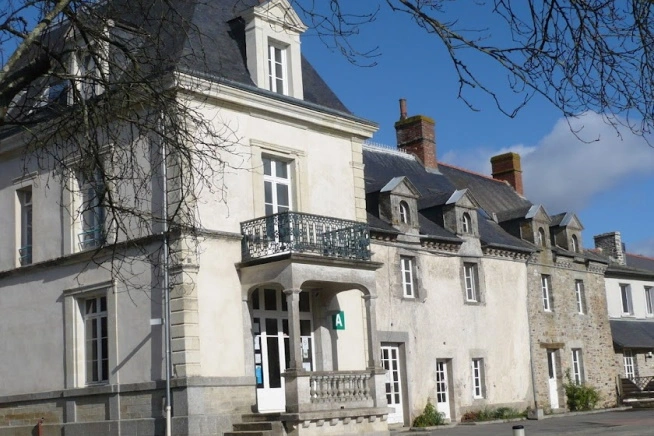 Bâtiment ancien en pierre et enduit clair composé de plusieurs corps de bâtiment reliés entre eux. La façade principale comprend un perron couvert avec balcon en ferronnerie, de hautes fenêtres blanches et une toiture en ardoise. Des branches d’arbres encadrent l’image au premier plan.