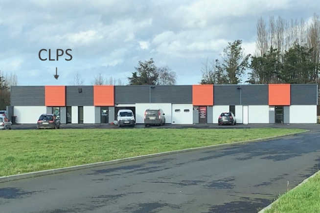 Bâtiment de plain-pied aux façades blanches et gris foncé, rythmé par plusieurs panneaux orange. Plusieurs véhicules sont stationnés devant le bâtiment, séparé de la route par une grande pelouse verte. Une flèche et le texte « CLPS » indiquent l’emplacement du centre.