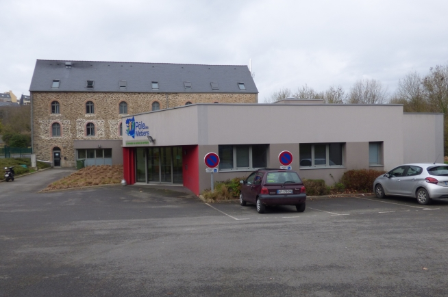 Bâtiment composé d’une partie contemporaine de plain-pied aux façades beige clair et rouge, reliée à un bâtiment en pierre de plusieurs niveaux avec toiture en ardoise. Plusieurs véhicules sont stationnés sur le parking situé au premier plan.