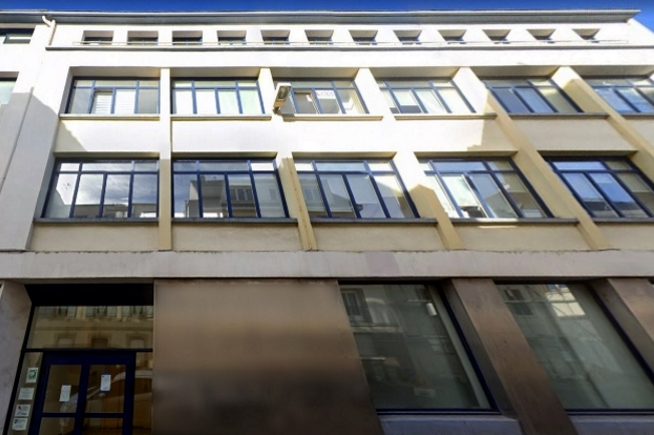 Bâtiment de plusieurs niveaux aux façades beige clair avec de grandes fenêtres alignées sur toute la hauteur de l’immeuble. L’entrée est située en rez-de-chaussée sous une façade vitrée et un encadrement sombre.