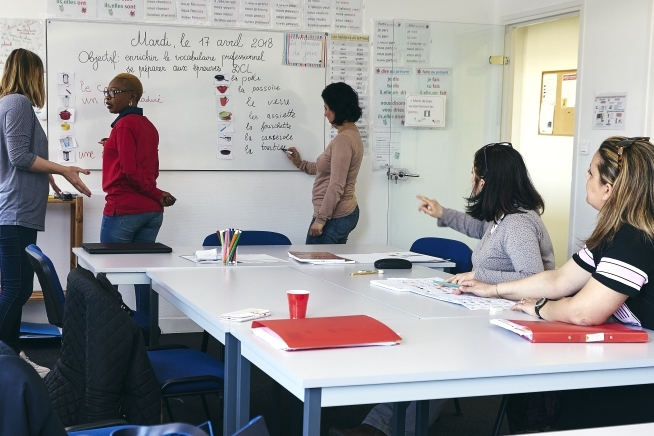 Cinq femmes participent à une activité de formation dans une salle de cours. Deux sont debout devant un tableau blanc couvert de mots, de phrases et d’illustrations, tandis que trois autres sont assises autour d’une table et suivent l’exercice à l’aide de supports pédagogiques.
