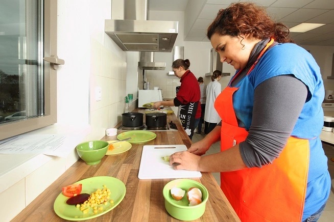 Femme portant un tablier orange et un haut bleu préparant des légumes sur une planche à découper dans une cuisine pédagogique. À l’arrière-plan, trois autres personnes travaillent sur des postes de cuisson et de préparation.
