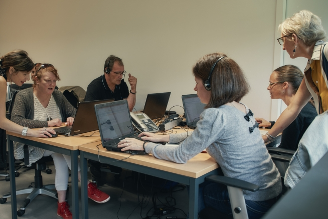 Six personnes sont réunies autour de plusieurs tables équipées d’ordinateurs portables. Toutes portent un casque audio avec micro et utilisent un ordinateur ou consultent l’écran d’un autre participant. Des téléphones, documents et câbles sont disposés au centre des tables.