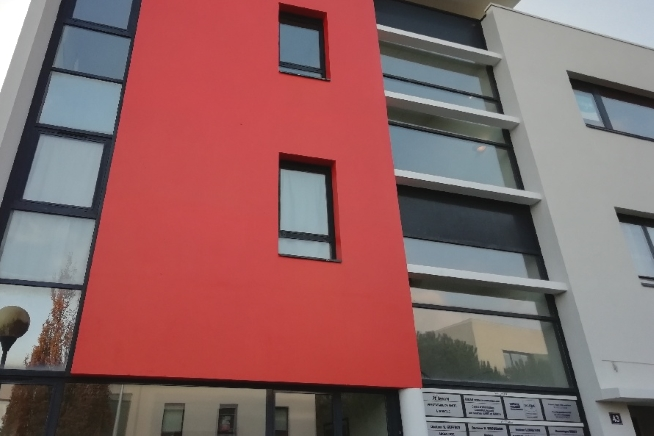 Bâtiment moderne de plusieurs niveaux avec une façade rouge vif encadrée par des parties blanches et grises. De grandes surfaces vitrées occupent le rez-de-chaussée et les étages, tandis que plusieurs panneaux d’information sont visibles près de l’entrée.