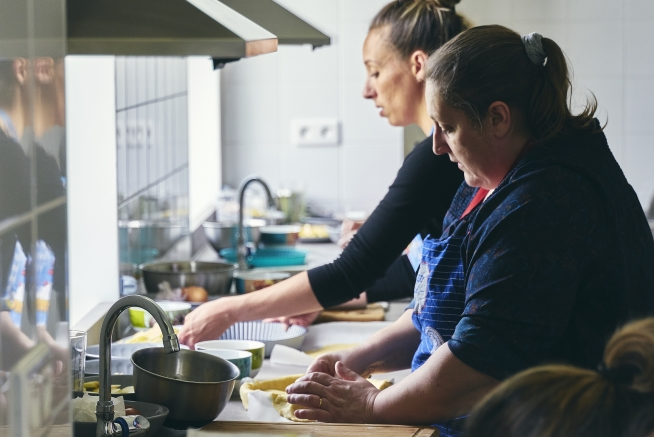 Deux femmes travaillent côte à côte dans une cuisine pédagogique. Au premier plan, l'une en tablier bleu prépare une pâte sur un plan de travail en bois, tandis que l'autre réalise une préparation culinaire à proximité. Un évier, des casseroles et du matériel de cuisine sont visibles autour des postes de travail.