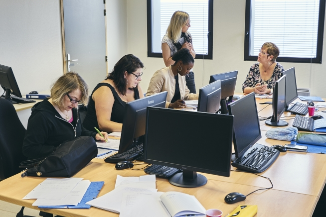 Cinq personnes participent à une séance de formation autour d’une grande table équipée de postes informatiques. Quatre personnes sont assises devant des écrans et des documents de travail, tandis qu’une formatrice se tient debout près des fenêtres au fond de la salle.