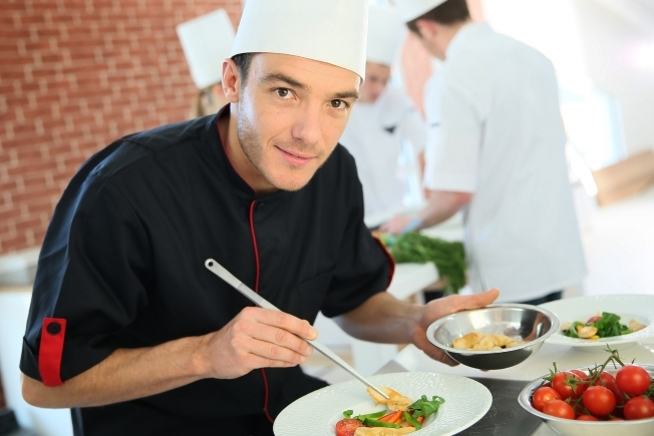Homme en tenue de cuisine tenant une assiette tout en dressant une préparation alimentaire. D'autres personnes en tenue professionnelle travaillent dans la cuisine à l'arrière-plan.