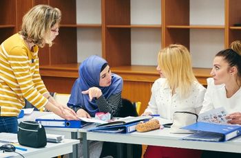 Quatre femmes réunies autour d'une table de travail couverte de classeurs, de pochettes et de documents. Une femme portant un haut à rayures se tient debout et échange avec trois autres femmes assises. Des étagères en bois vides occupent l'arrière-plan de la salle.