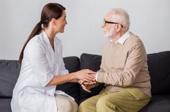 Femme en tenue professionnelle assise face à un homme âgé, tenant ses mains sur un canapé.
