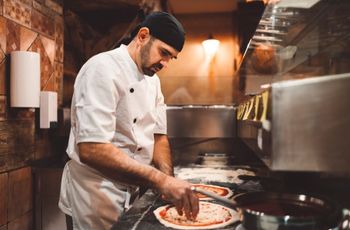 Homme en tenue de cuisine garnissant une pâte à pizza sur un plan de travail dans une cuisine professionnelle.
