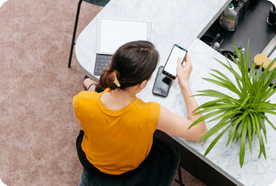 Femme assise à un bureau consultant un téléphone mobile. Un ordinateur portable et une plante sont visibles sur le plan de travail.