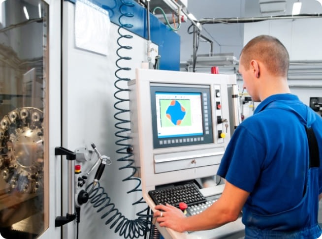 Homme en tenue de travail bleue utilisant un pupitre de commande relié à une machine industrielle. Un écran de contrôle, un clavier et plusieurs équipements automatisés sont visibles dans l'atelier.