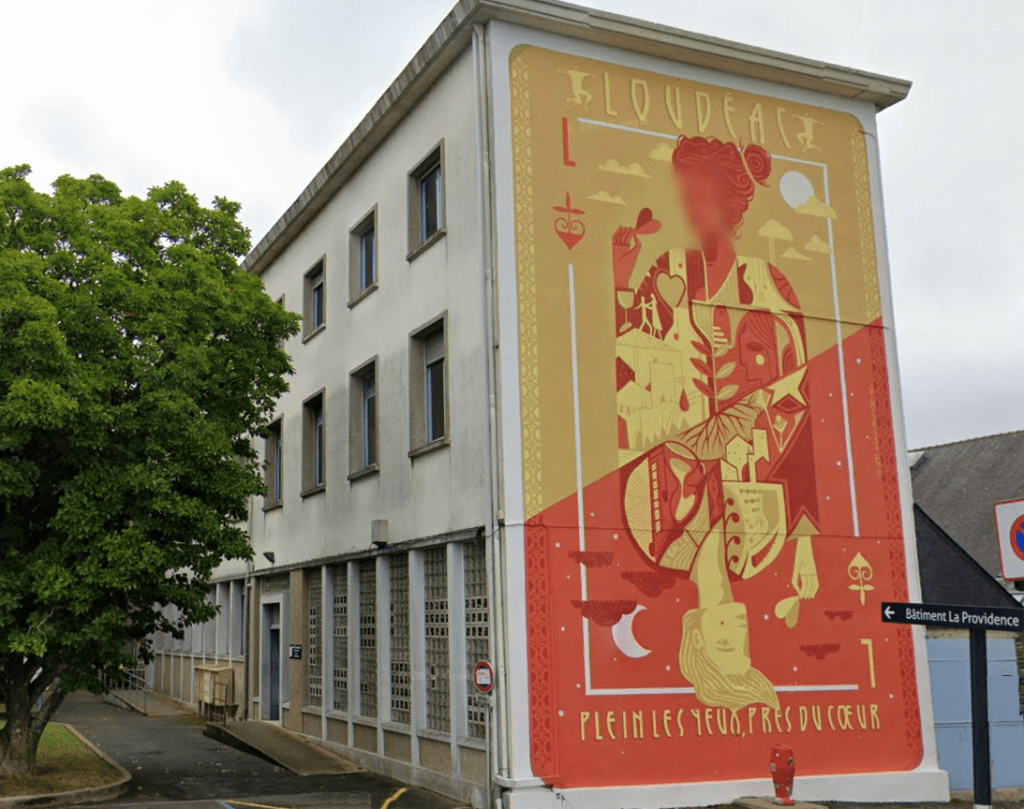 Bâtiment clair de trois niveaux dont le pignon est recouvert d’une grande fresque rouge et beige portant l’inscription « Loudéac », à proximité d’un arbre et d’une voie piétonne.