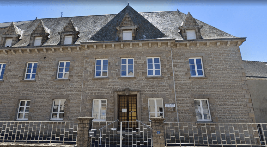 Grand bâtiment en pierre grise à trois niveaux avec toiture en ardoise, lucarnes, porte centrale et fenêtres symétriques en façade.