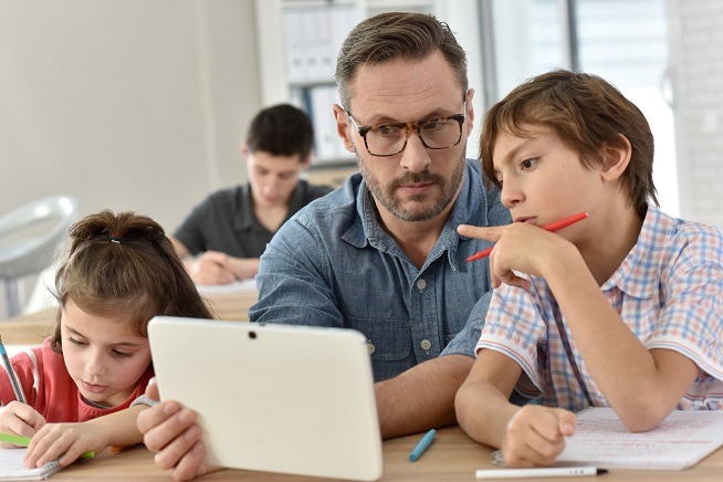 Homme assis avec deux enfants autour d'une tablette numérique dans une salle de classe ou d'activité.