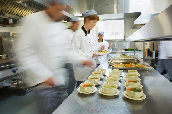 Personnes en tenue professionnelle de restauration préparant et distribuant des repas dans une cuisine collective. Plusieurs bols de soupe sont alignés sur un plan de travail en inox.