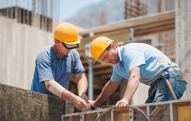 Deux hommes portant des casques de chantier jaunes manipulent des armatures métalliques sur un chantier de construction. Des coffrages et structures en béton sont visibles à proximité.