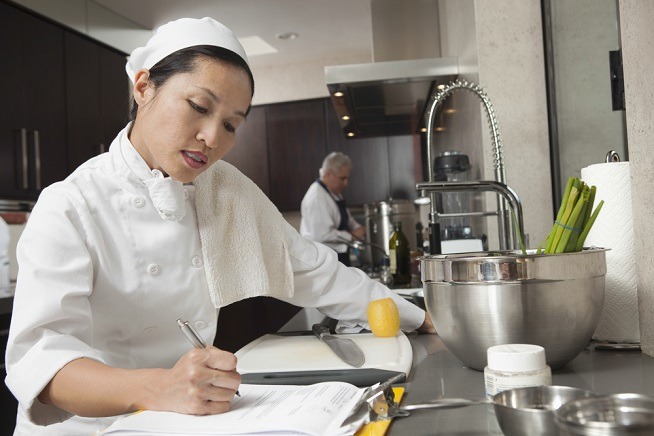 Femme en tenue professionnelle de cuisine écrivant sur un document dans une cuisine. Des ingrédients, des ustensiles et un plan de travail sont visibles à proximité.