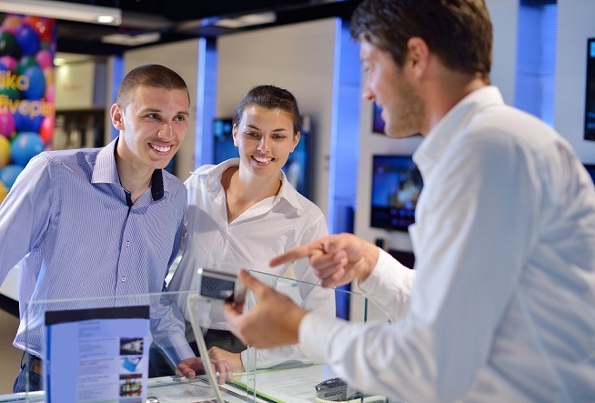 Trois personnes échangent dans un magasin spécialisé en produits électroniques. Un homme présente un produit tandis que deux autres se tiennent derrière un comptoir d'exposition.