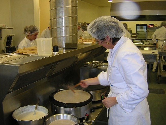 Femme en tenue professionnelle de cuisine et charlotte préparant une crêpe sur une billig dans un laboratoire de cuisine.