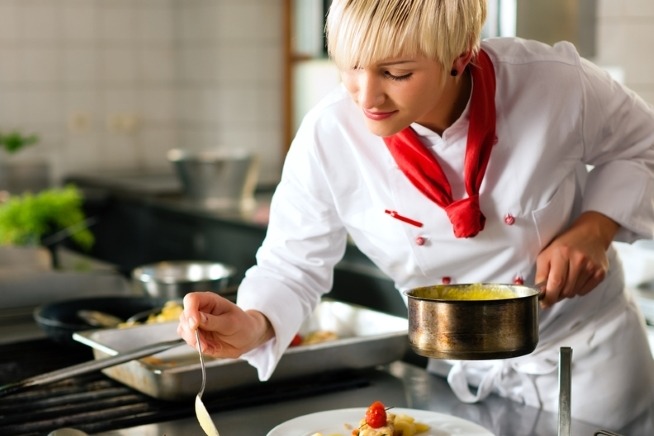 Femme en tenue de cuisine blanche tenant une casserole et dressant une préparation dans une assiette dans une cuisine professionnelle.