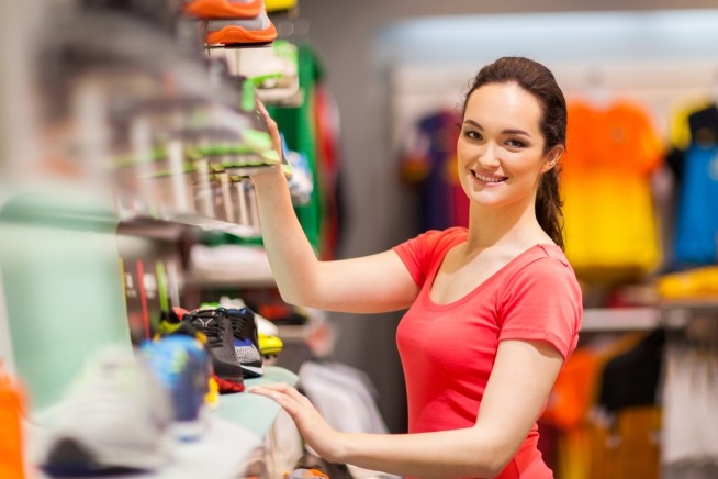 Image montrant une femme dans un magasin, debout devant un rayon de chaussures et d'articles de sport. Plusieurs produits sont exposés sur les étagères au premier plan et en arrière-plan.