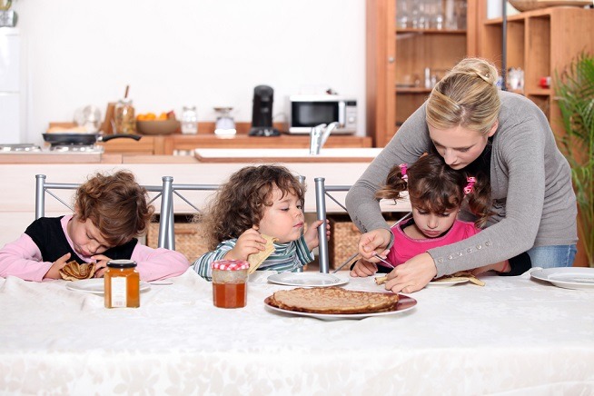 Image montrant trois jeunes enfants assis autour d'une table dans une cuisine. Une femme se penche vers l'un des enfants pour l'aider à étaler quelque chose sur sa crêpe. Des pots de confiture et des crêpes sont posés sur la table.