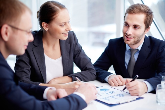 Trois personnes en tenue professionnelle échangent autour de documents et de graphiques lors d'une réunion de travail.