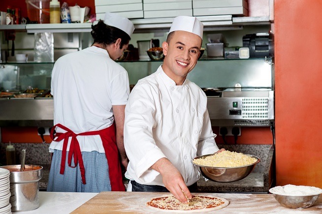 Homme en tenue professionnelle de cuisine préparant une pizza sur un plan de travail. Un autre homme travaille dans une cuisine professionnelle à l'arrière-plan.