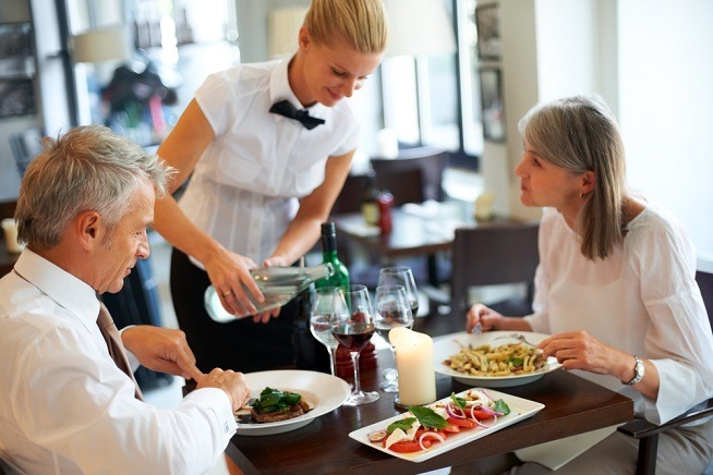 Femme en tenue de service versant de l'eau à une table occupée par deux clients dans un espace de restauration. Des assiettes et des verres sont disposés sur la table.